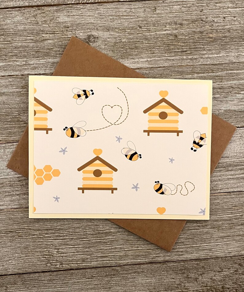 Puede incluir: Una tarjeta amarilla con fondo blanco y un patr&oacute;n de colmenas, abejas y corazones. La tarjeta tiene un sobre marr&oacute;n claro.