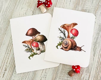 May include: Two blank greeting cards with watercolor illustrations of mushrooms and a snail. The card on the left features a cluster of mushrooms with red and white caps. The card on the right features a snail with a brown shell and a red and white mushroom.