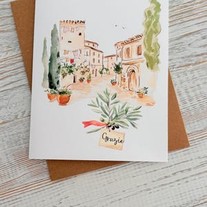 May include: Watercolor greeting card featuring a European village scene with buildings and a cobblestone street. An olive branch with the word "Grazie" is in the foreground. The card is on a brown envelope.