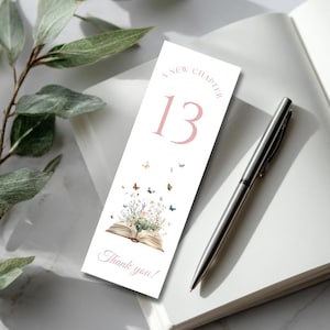 May include: A white bookmark with the text "A New Chapter" and "13" in pink. The bookmark features an open book with flowers and butterflies, and the words "Thank you!" are at the bottom. A silver pen and a notebook are also in the image.