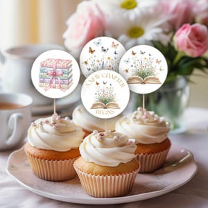 Peut inclure: Des cupcakes avec un glaçage blanc et des décorations. Les décorations présentent des illustrations de livres, de fleurs et de papillons. L'une des décorations indique "A New Chapter Begins". Les cupcakes sont sur une assiette rose.