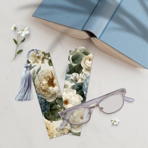 May include: Two floral bookmarks with a light blue tassel, a pair of glasses, and a book. The bookmarks feature a cream background with white and beige roses, blue hydrangeas, and green leaves. The glasses have a light purple frame.