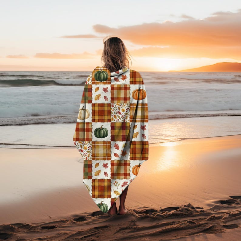 May include: A hooded blanket with a patchwork design featuring pumpkins, leaves, and plaid squares in shades of orange, green, and brown. The blanket is draped over a person standing on a beach at sunset.