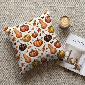 May include: Square decorative pillow with a cream background featuring a watercolor pattern of pumpkins, gourds, and fall leaves in shades of orange, brown, and yellow. A cup of coffee and a magazine are next to the pillow.