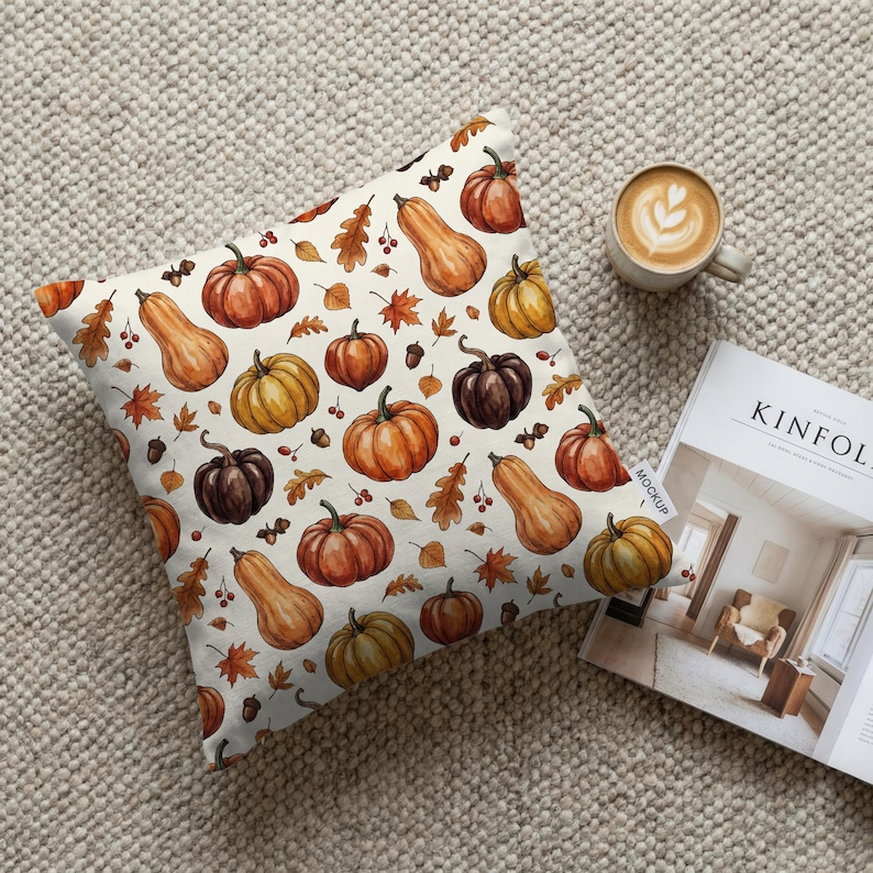 May include: Square decorative pillow with a cream background featuring a watercolor pattern of pumpkins, gourds, and fall leaves in shades of orange, brown, and yellow. A cup of coffee and a magazine are next to the pillow.