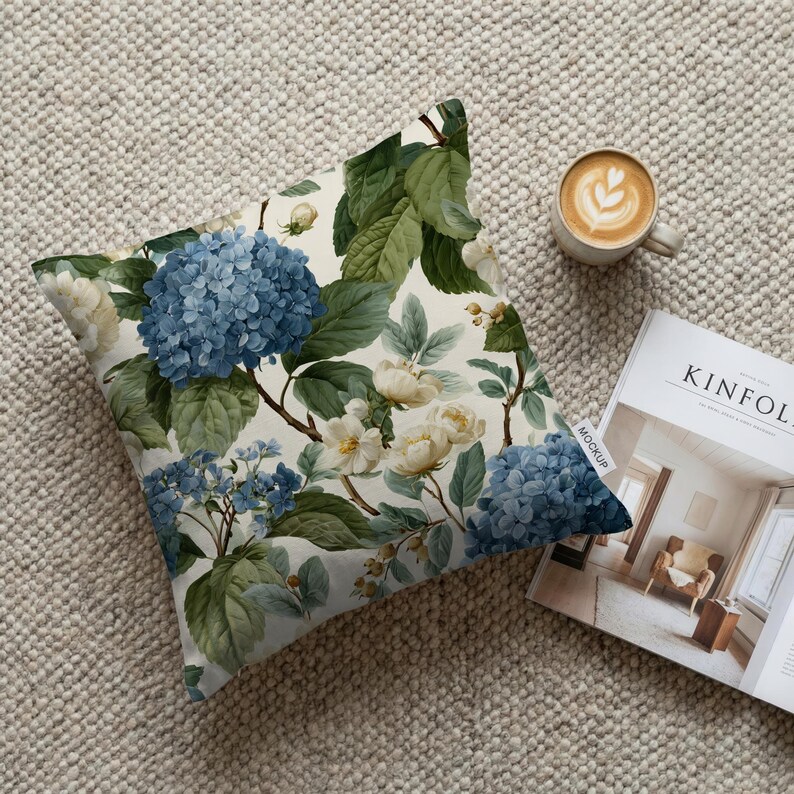 May include: Square throw pillow with a floral pattern featuring blue hydrangeas and white flowers on a cream background. The pillow is next to a cup of coffee and a magazine.