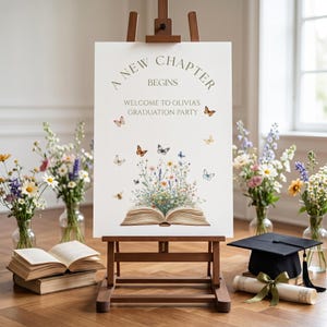 May include: A graduation party sign on a wooden easel. The sign reads "A New Chapter Begins" and "Welcome to Olivia's Graduation Party." Butterflies and flowers decorate the sign. A graduation cap and diploma are on the floor.