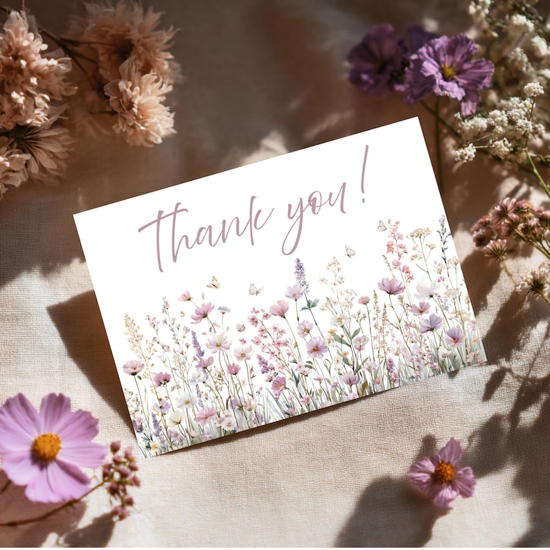 May include: A white card with the words "Thank you!" in a soft purple script. The bottom of the card features a watercolour illustration of wildflowers in shades of pink, purple, and white, with small butterflies.