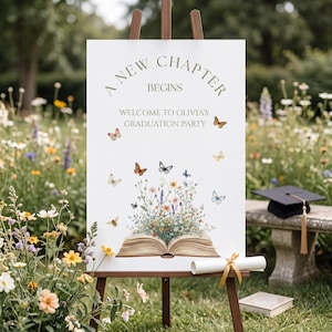 May include: A white graduation party sign on a wooden easel. The sign reads "A New Chapter Begins" and "Welcome to Olivia's Graduation Party." Butterflies and flowers decorate the sign. A graduation cap and diploma are nearby.