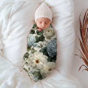 May include: A newborn baby swaddled in a floral print blanket, with a matching white knit cap. The blanket features large white, blue, and green flowers. The baby is lying on a white sheet.