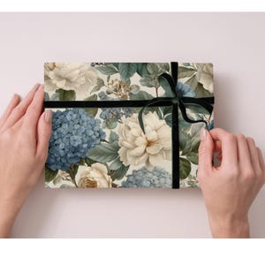 May include: A floral gift box featuring a pattern of blue hydrangeas, cream-colored roses, and green foliage. The box is wrapped with a black velvet ribbon tied in a bow. The box is held by two hands.