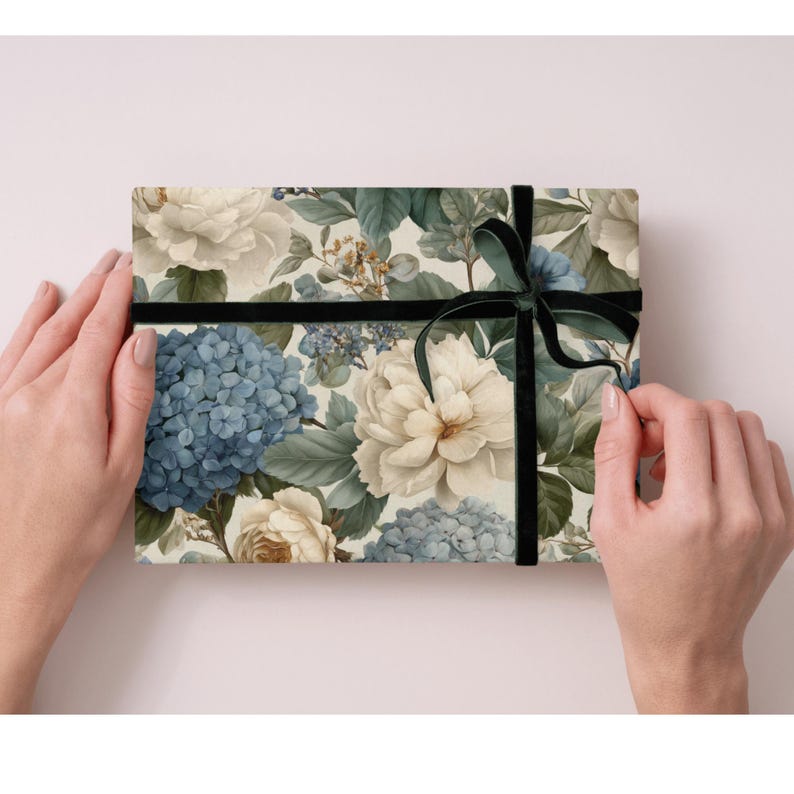 May include: A floral gift box featuring a pattern of blue hydrangeas, cream-colored roses, and green foliage. The box is wrapped with a black velvet ribbon tied in a bow. The box is held by two hands.