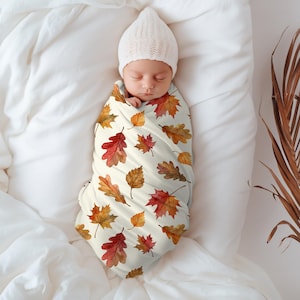 May include: A newborn baby wrapped in a cream-colored swaddle blanket adorned with watercolor autumn leaves in shades of red, orange, and brown. The baby wears a white knit cap and rests on a white sheet.