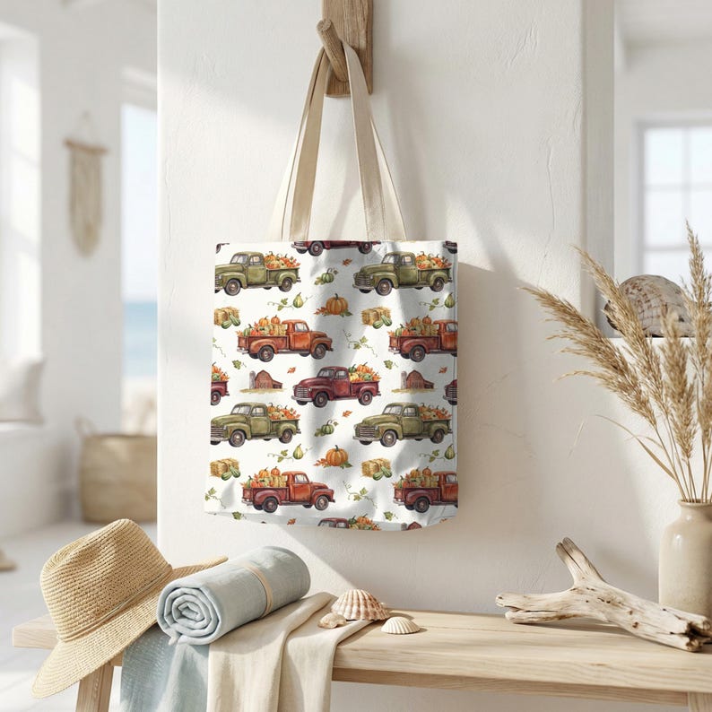 May include: A cream-colored tote bag featuring a repeating pattern of vintage trucks filled with pumpkins, gourds, and hay bales. The trucks are in shades of green, red, and orange, set against a white background. The bag has natural-colored handles.