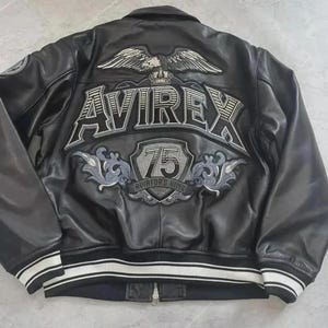 May include: A black leather jacket featuring a large embroidered graphic on the back. The design includes an eagle, the text AVIREX, a shield with the number 75, and the words AVIATOR'S KING. The jacket has a ribbed hem and cuffs with white stripes.