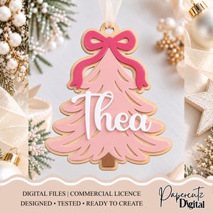 May include: A layered wooden Christmas tree ornament in shades of pink, featuring a dark pink bow at the top and the name Thea in white script lettering across the center. The ornament hangs from a light-colored ribbon loop.