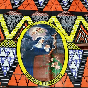 May include: A colorful fabric with a black background and a yellow circle in the center. The circle contains a religious image of a woman praying with a cross in the background. The text around the circle reads "Mtakatifu Rita Wa Cascia Utuombee Sisi Wakosefu".