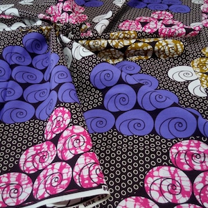 Può includere: Un tessuto piegato con un motivo ripetuto di spirali viola, rosa, bianche e dorate su uno sfondo marrone scuro. Il tessuto presenta un piccolo motivo a pois bianchi. Il testo "POWER BLOCK WAX 323" è visibile.