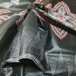 Può includere: Tessuto nero con un motivo geometrico bianco e rosso. Il tessuto ha una finitura lucida ed è piegato a metà. Il testo "SUPER AVOCAN" è visibile sul tessuto.