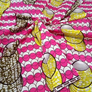 以下が含まれることがあります： ピンクの円と黄色と茶色の曲線模様が繰り返される白い生地。生地の下端に「Super Power Block Wax」という文字が印刷されています。