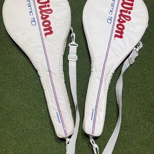 May include: Two white Wilson Ceramic 110 tennis racket covers with red and blue accents. Each cover features the Wilson logo in red, the text Ceramic 110 in blue, and the word Largehead in blue near the handle, complete with a white shoulder strap.