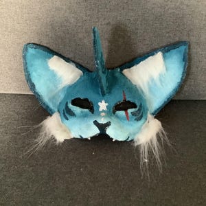 Può includere: Una maschera animale blu e bianca con grandi orecchie a punta, una protuberanza centrale a forma di pinna, fori neri per gli occhi, una stella bianca sulla fronte, una linea rossa verticale sopra l'occhio sinistro e accenti di pelliccia sintetica.