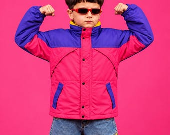 Pode incluir: Um casaco com blocos de cores, apresentando um corpo rosa brilhante com mangas e painéis superiores no peito em roxo escuro. O casaco tem uma gola alta com forro interior amarelo, fecho frontal com botões e dois bolsos laterais, usado com calças de ganga azuis.