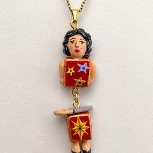 Puede incluir: Un collar con colgante que muestra una figura de cabello negro, con un torso cuadrado rojo decorado con estrellas amarillas y moradas, y una sección inferior cuadrada roja con una estrella amarilla grande. La figura tiene una pequeña bandeja gris y lleva zapatos negros.