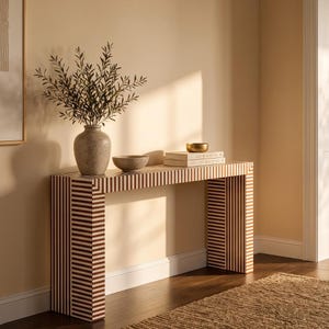 May include: A rectangular console table featuring a brown and white vertical striped pattern. On top sits a textured beige vase with green leafy branches, a small gray bowl, and a stack of two white books topped with a small gold bowl.