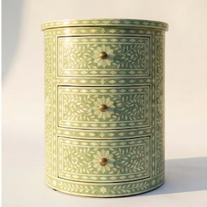 May include: A cylindrical three-drawer chest featuring a light green finish with intricate white floral and leaf inlay patterns. Each drawer is equipped with a small, round, metallic knob centered on a detailed botanical design.