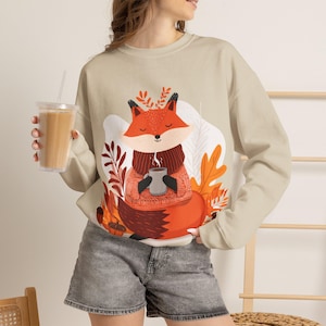 Peut inclure: Un sweat-shirt beige à col rond orné d'une illustration graphique représentant un renard portant un pull et une écharpe, tenant une tasse fumante. Le renard est entouré de feuilles d'automne orange et rouges et de glands sur un fond blanc.