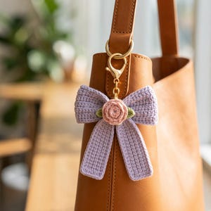 May include: A lavender crochet bow bag charm featuring a central pink crocheted rose with two small green leaves. The bow is attached to a gold-toned metal lobster clasp and chain, which is clipped onto the handle of a tan leather handbag.