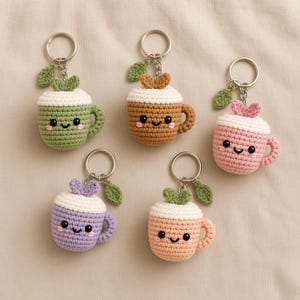Puede incluir: Cinco llaveros de ganchillo con forma de pequeñas tazas con caras sonrientes. Cada taza tiene una parte superior blanca, un asa y una pequeña hoja verde unida al anillo de metal plateado. Las tazas son verdes, marrones, rosas, moradas y naranja.