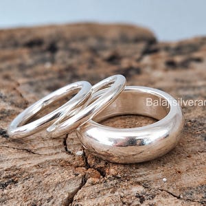 Puede incluir: Tres anillos de plata pulida de diferentes grosores dispuestos juntos. El conjunto incluye dos bandas finas y lisas y una banda más ancha y redondeada. El texto Balajisilverarts26IN es visible en el lado derecho cerca de los anillos.