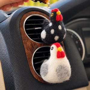 Può includere: Due clip deodoranti per auto a forma di gallina in lana infeltrita fissate a una bocchetta dell'aria. Una gallina è nera a pois bianchi, l'altra è bianca con ali grigie. Entrambe hanno creste rosse e becchi gialli. Si vede un flacone di olio.