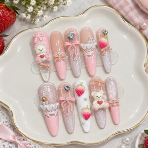 Peut inclure: Un ensemble de dix faux ongles avec une palette de couleurs rose et blanc. Les motifs incluent des breloques oursons en 3D, des appliques fraises, des finitions pailletées, des motifs de dentelle blanche, des petits nœuds et des strass en cœur.