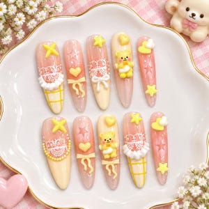 Peut inclure: Un ensemble de dix faux ongles avec une palette de couleurs rose et crème. Les motifs incluent des oursons jaunes en 3D, de la dentelle blanche, des étoiles jaunes, des cœurs, des nœuds et des quadrillages. Finition brillante et forme amande.