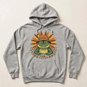 May include: A heather grey hooded sweatshirt featuring a graphic print of a green alligator wearing an orange patterned bucket hat. The alligator is centred in front of a sunburst design with small mushrooms and flowers surrounding the base.