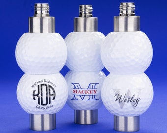 May include: Three personalized golf ball bottle stoppers. The first stopper has a monogram with the initials "KDA" and the text "Kathrina Dobrusin 06.26.1990". The second stopper has the text "MACKEY" with a monogram of the letter "M". The third stopper has the name "Wesley" written on it.