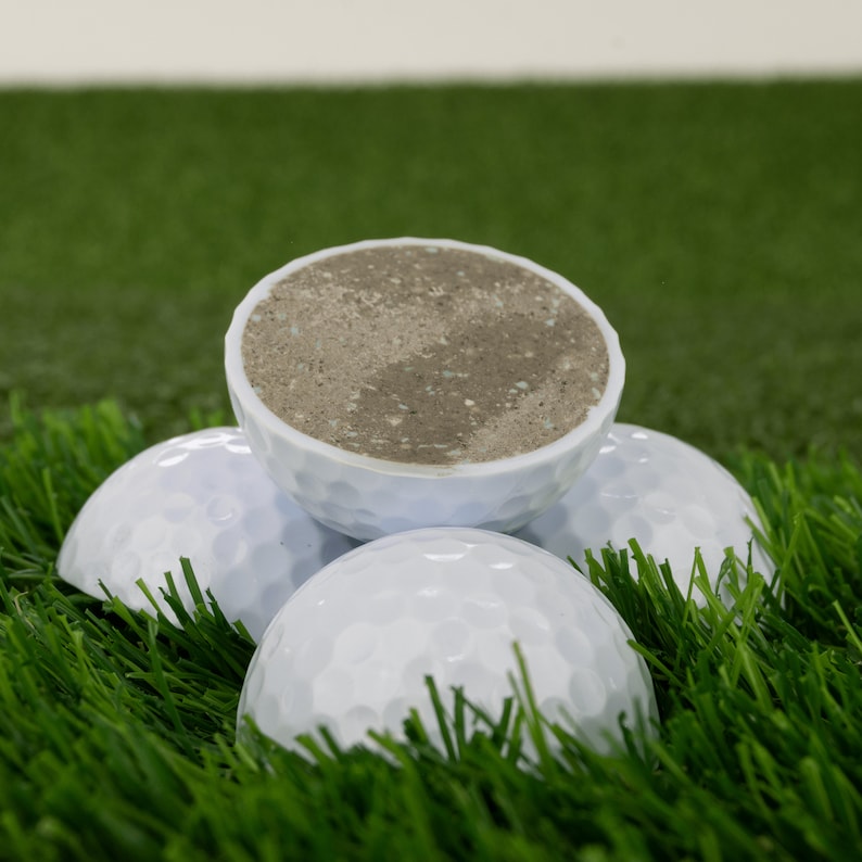 May include: Four white golf balls, one of which is cut open to reveal a brown and gray interior. The golf balls are sitting on a green artificial grass surface.