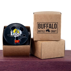 May include: A black and white illustration of Mickey Mouse with red shorts and yellow shoes on a black hockey puck. The puck is in a brown cardboard box with the words "BUFFALO BOTTLE CRAFT" printed on the box.