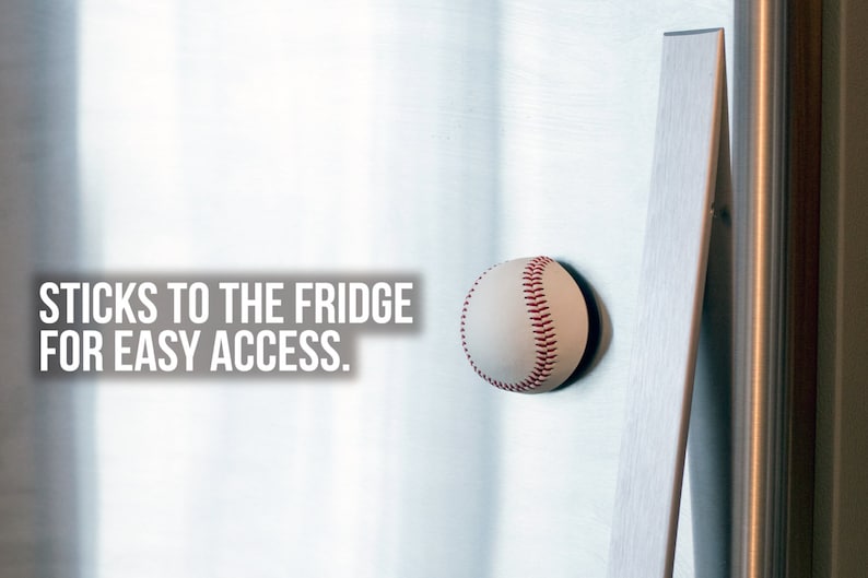 May include: A white baseball with red stitching is attached to a silver refrigerator with a magnet. The text "STICKS TO THE FRIDGE FOR EASY ACCESS." is visible in the image.