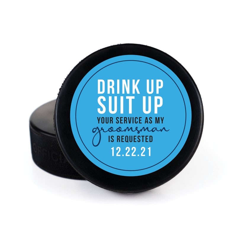 May include: A black hockey puck with a blue circle sticker that says "Drink Up Suit Up Your Service As My Groomsman Is Requested 12.22.21"
