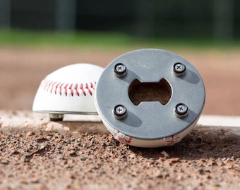 May include: A baseball-shaped bottle opener with a metal top and a baseball stitching design. The bottle opener is sitting on a dirt surface.