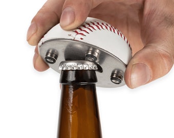 May include: A white baseball-shaped bottle opener with red stitching is being used to open a brown glass bottle. The bottle opener has a silver metal base with four magnets.