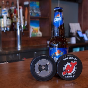May include: Three black hockey puck bottle openers, one with a metal bottle opener, one with the New Jersey Devils logo, and one with the text "The Puck Stops Here". A bottle of Labatt Blue beer is in the background.