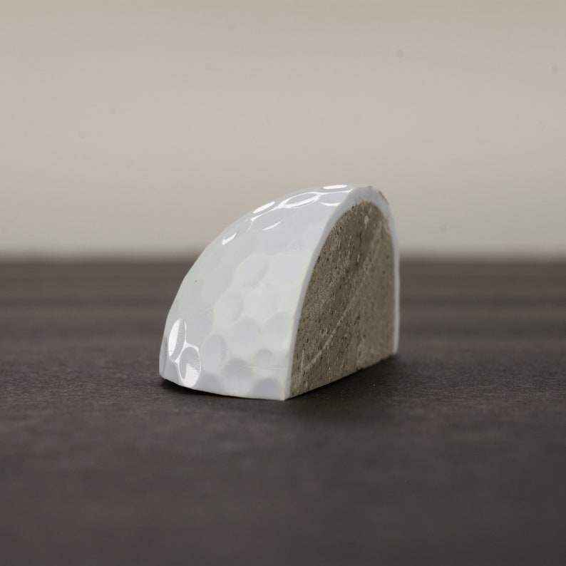 May include: Half of a white ceramic golf ball with a textured surface. The other half of the ball is made of concrete.