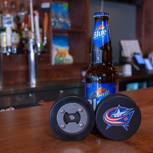 以下が含まれることがあります： 「The Puck Opener」という文字と銀色の栓抜きが付いた黒いホッケーパックの栓抜き。栓抜きの隣には、白い星と「CBJ」という文字が書かれた青と赤のホッケーパックがあります。ホッケーパックの後ろには、ラバッツブルービールの瓶があります。