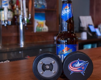 May include: A black hockey puck bottle opener with the text "The Puck Opener" and a silver bottle opener. A blue and red hockey puck with a white star and the text "CBJ" is next to the bottle opener. A bottle of Labatt Blue beer is behind the hockey pucks.