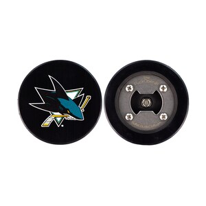 以下が含まれることがあります： 片側にサンノゼ・シャークスのロゴ、もう片側に「The Puck Opener」の文字と栓抜きがついた、黒と白のホッケーパック栓抜きです。